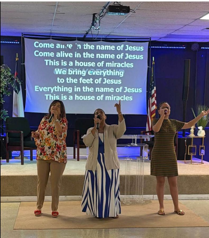 Three women singing with microphones on stage in a church sanctuary with religious text displayed on a screen behind them