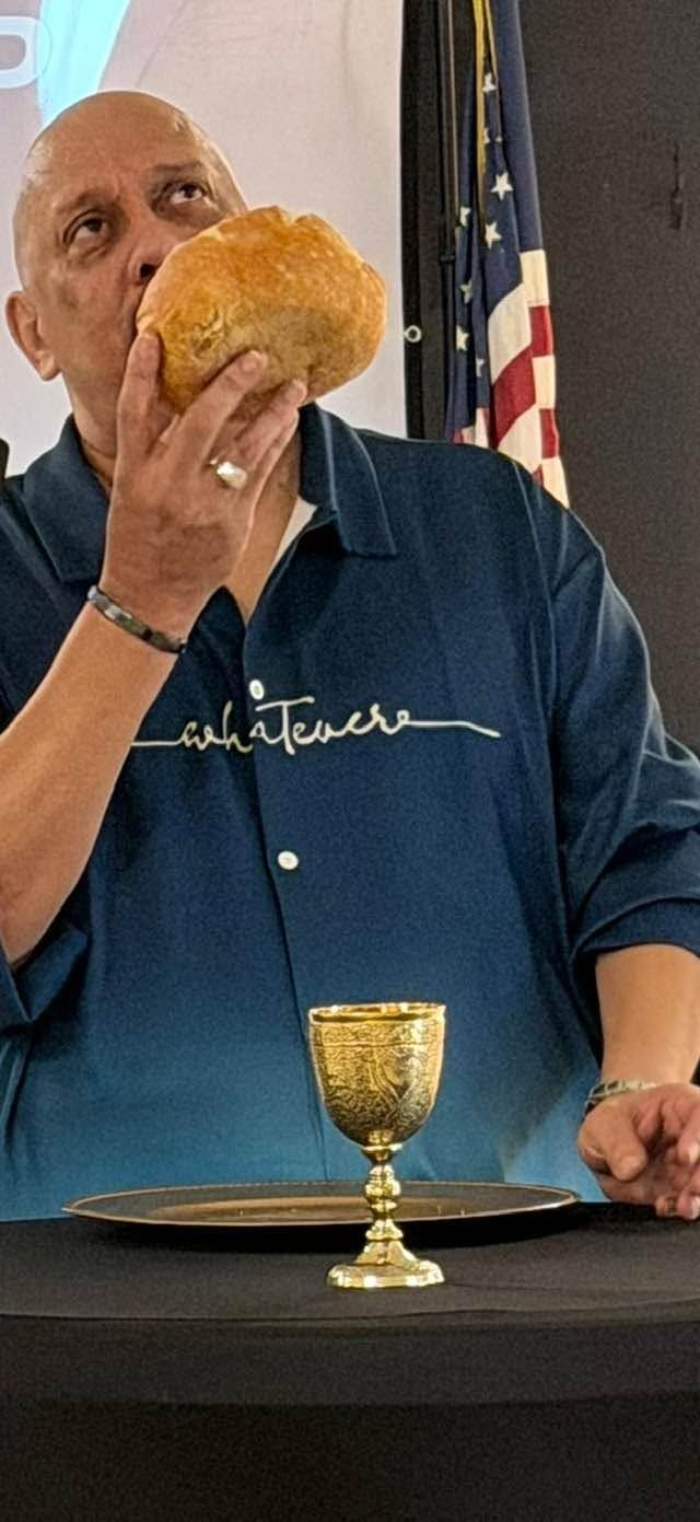 Man in blue polo shirt holding bread near golden chalice with American flag in background