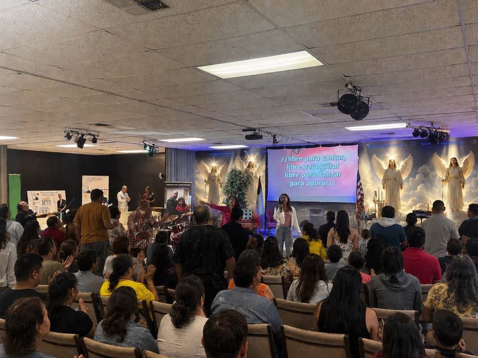 Indoor religious service with large congregation seated facing stage with projection screen and angelic imagery