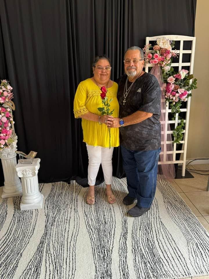 Man and woman posing together holding a red rose in front of floral backdrop