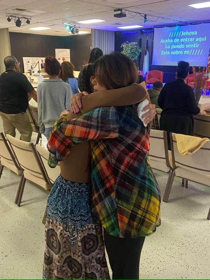 Two women embrace from behind in a community meeting room with blue lighting and a projection screen displaying Spanish text